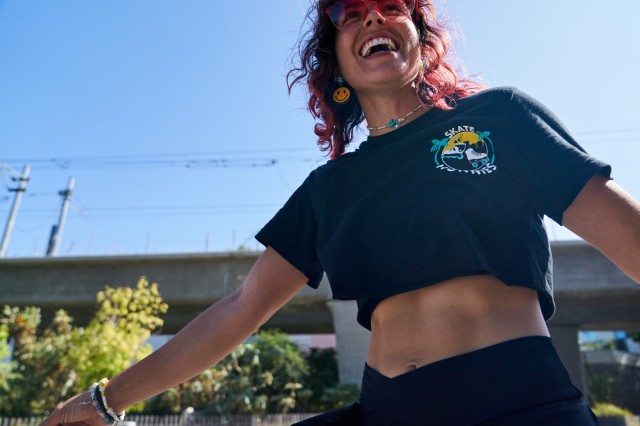 Woman with long pink hair laughing out loud, wearing a dark skirt and crop top with the logo "Skate Hunnies" on it.